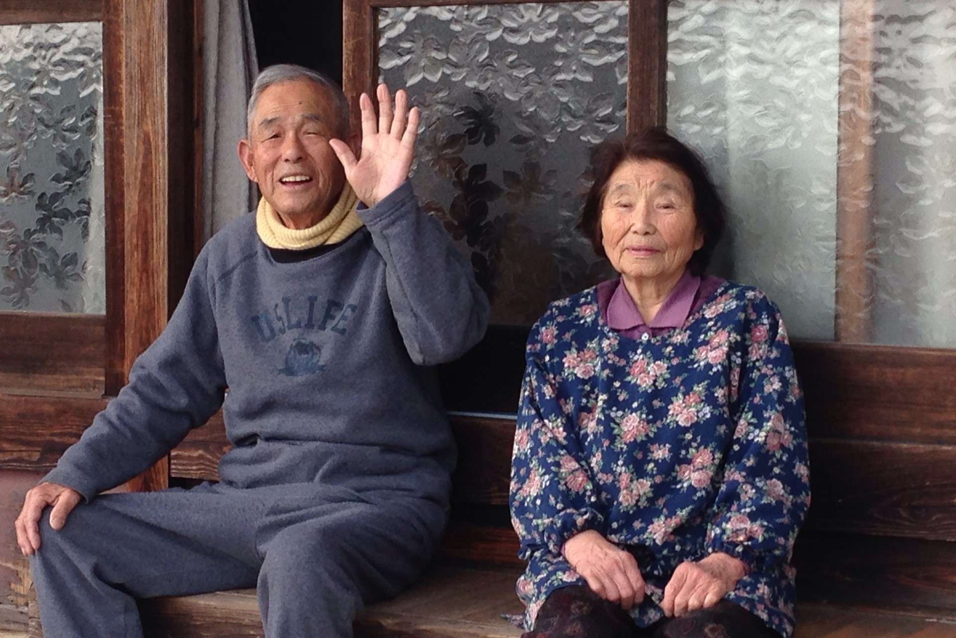 An elderly couple sitting on the porch of their house smiling and waving at us.