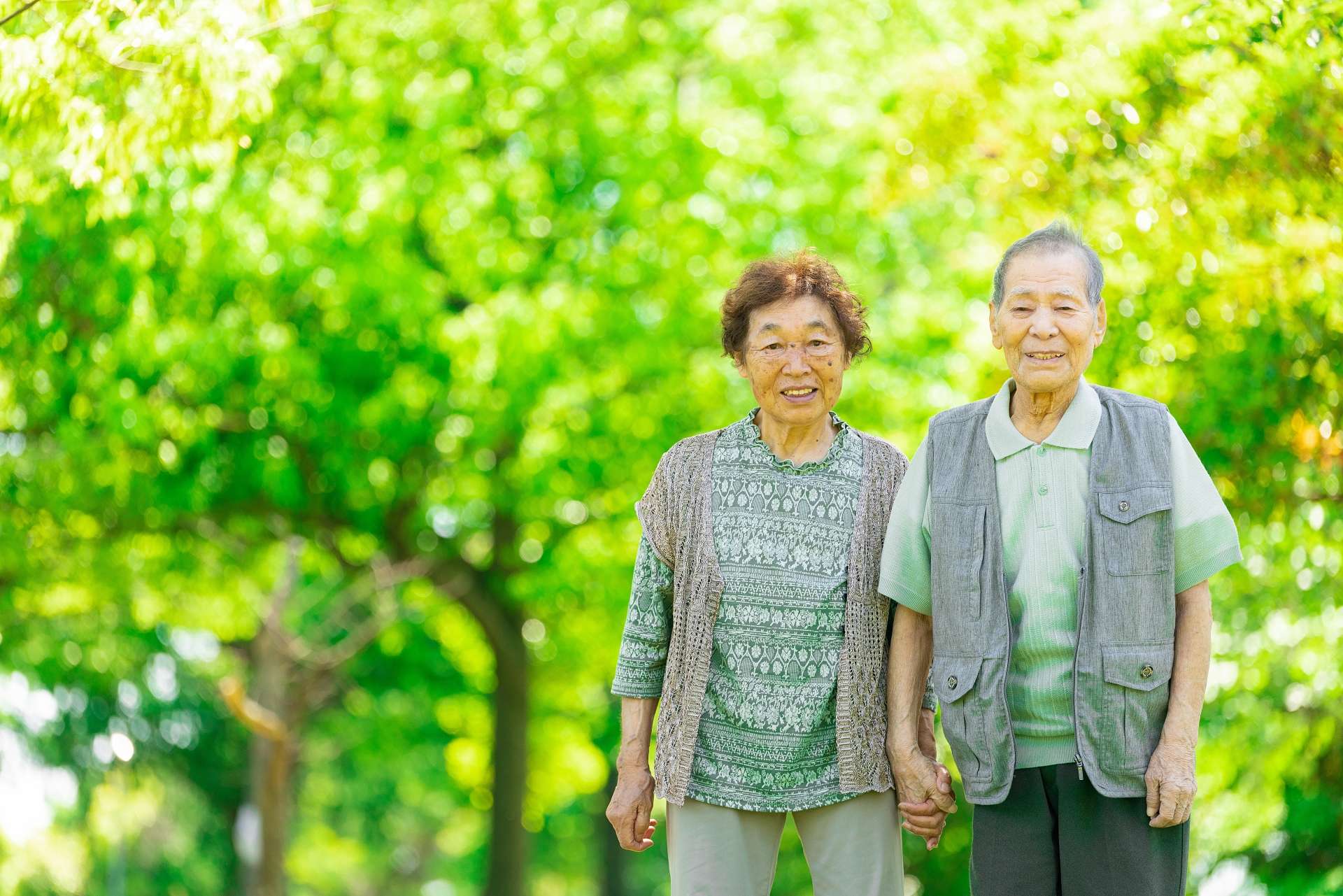 An elderly couple is smiling and taking a walk while holding hands in the sunlight filtering through the trees.